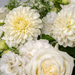 A close-up of a white floral arrangement featuring large dahlia-like blooms, roses, and green foliage on a light gray background.