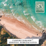 Aerial beach scene with turquoise ocean waves, pale sand, dark rocky shoreline, and a postage stamp graphic in the top right reading Australia Viola Floral. Text at the bottom reads Passport to Bloom Byron Bay - Bubbles & Natives.